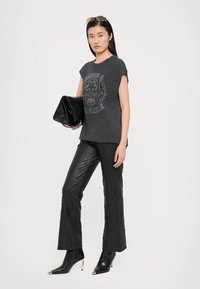 Grijze grafische t-shirt met schedelontwerp, gecombineerd met zwarte textuurbroek met flared pijpen en zwarte enkelaars met puntige neuzen en gouden accenten.