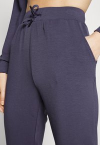 Gros plan d'une personne portant un pantalon à cordon de serrage de couleur violet foncé avec une taille élastique et une poche latérale, la main reposant dans la poche.