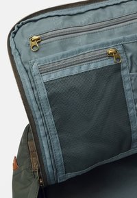 Intérieur d'un sac en tissu vert avec deux compartiments à fermeture éclair, l'un avec un panneau en maille, et une doublure lisse gris clair. Accents de ferrures dorées.