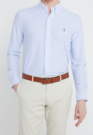Homme portant une chemise à rayures bleu clair et blanc boutonnée, rentrée dans un pantalon beige avec une ceinture en cuir marron, main droite dans la poche.