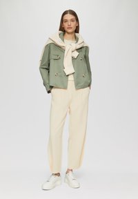Veste verte à double boutonnage, pull crème noué au cou, pantalon beige clair et baskets blanches, sur fond uni.