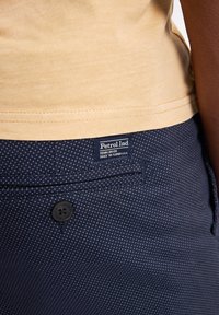 Pantaloni cu model marinăresc, cu buline albe, având un nasture negru și o etichetă cu inscripția "Petrol Ind." Cămașă superioară de culoare bej vizibilă.