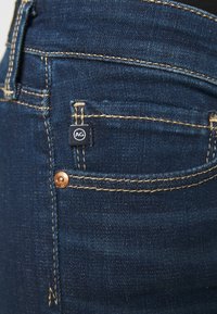 Tmavě modré džíny z denimu mají zlaté prošívání, malou štítkovou nášivku AG a měděné nýty na zadní kapse, což umocňuje klasický design.