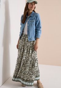 Femme portant une casquette bleu clair, une veste en jean, une chemise blanche, une jupe longue à motifs et des chaussures beiges à enfiler, debout contre un mur pâle.