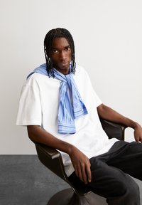 Weißes, übergroßes T-Shirt mit einem blauen gestreiften Schal um den Hals; sitzt in einem dunklen, glatten Stuhl vor einem neutralen Hintergrund.