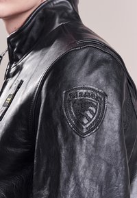 Veste en cuir noir avec un col haut ; présente un patch sur l'épaule gauche, une texture lisse et des coutures détaillées le long des coutures.