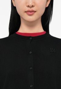 Kadar u žarištu osobe koja nosi crni kardigan s dugmadi preko crvene majice, s vidljivim izvezenim "HUGO" logom na lijevoj strani grudi.