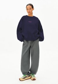 Übergroßer marineblauer Pullover mit lila Schriftzug, kombiniert mit weiten grauen Hosen und mehrfarbigen Turnschuhen mit grünen Schnürsenkeln und strukturierten Sohlen.