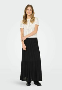 Top corto de punto blanco con un patrón texturizado, combinado con una larga falda maxi negra de volantes. La modelo lleva botas de tobillo.