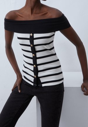 Persoon draagt een zwart-wit gestreept topje met blote schouders en gouden knopen, gecombineerd met een zwarte broek, staand tegen een grijze achtergrond.