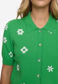 Zelený krátký kardigan s vroubkovaným límcem, zdobený bílými květinovými vzory a kulatými knoflíky po celé přední straně.