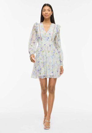 Femme portant une robe à manches longues à col en V, fleurie, avec des volants et des détails en dentelle, associée à des talons ouverts argentés, debout devant un fond blanc.