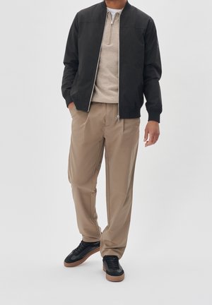 Man draagt een zwarte bomberjas, beige trui met rits, beige broek en zwarte sneakers met gumzolen, staand tegen een effen achtergrond.