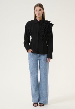Black shirt with ruffled detail on one shoulder, paired with light blue wide-leg trousers featuring vertical stripes and open-toe black shoes.