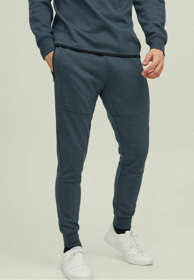 Grå joggingsbyxor i ett mjukt, elastiskt material. Har elastiska muddar, sidofickor och sömdetaljer för att ge extra visuell intress. 