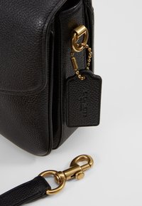 Coin de sac en cuir noir grainé avec garniture dorée et crochet de sangle amovible, doté d'une étiquette de marque en cuir sur une chaîne.