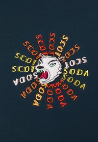 Testa di animale ringhiante ricamata circondata dalla ripetizione della parola "SCOT SODA" in fili rossi, arancioni e gialli su un tessuto scuro.