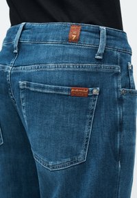 Πίσω θέα μπλε τζιν με καφέ ετικέτες μάρκας στη μέση και στην πίσω τσέπη, φορεμένα με μαύρο τοπ.