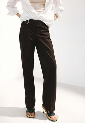 Mujer con pantalones negros de pierna ancha, camisa blanca sobre un top beige y sandalias negras de tacón alto, de pie con las manos en las caderas.