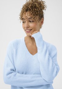 Göndör hajú nő világoskék V-nyakú pulóverben, mosolyogva lefelé néz, egyik kezét az állához közel tartva, egyszínű háttér előtt.