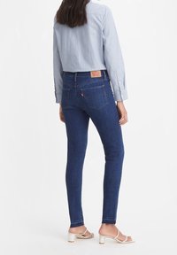 Vaqueros skinny de color azul oscuro con una cintura alta, que presentan bolsillos traseros y una etiqueta de cuero de la marca. Llevados con una camisa de botones color azul claro y sandalias.