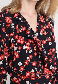 Musta kukallinen paita, jossa on pieniä punaisia ja valkoisia kukkia, röyhelökaulus ja kietaisumalli, valmistettu kevyestä kankaasta.