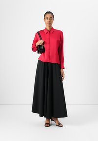 Chemise rouge à boutons avec col, associée à une jupe maxi noire fluide. L'ensemble est complété par des escarpins strappy noirs et un petit sac à main.