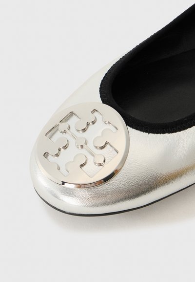 Ballerines en cuir argenté avec un bandeau élastique noir et un accent logo métallique rond sur le bout. Texture lisse et forme arrondie.
