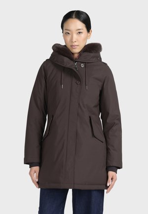 Femme portant un manteau d'hiver matelassé marron foncé avec une capuche doublée de fourrure, des boutons sur le devant et des poches zippées, debout devant un fond uni.
