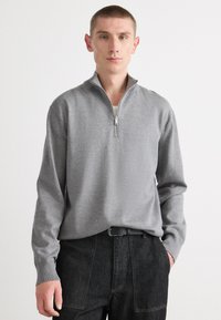 Grauer Reißverschluss-Pullover mit langen Ärmeln, gerippten Bündchen und flacher Strickstruktur, kombiniert mit dunklen Jeans, die einen Gürtel haben.