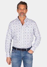 Witte blouse met lange mouwen en een blauw geometrisch patroon, met een knoopkraag en een logo op de borst; gecombineerd met lichtblauwe jeans en een donker gewatteerde riem.