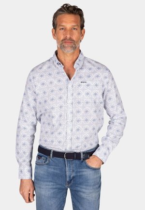 Witte blouse met lange mouwen en een blauw geometrisch patroon, met een knoopkraag en een logo op de borst; gecombineerd met lichtblauwe jeans en een donker gewatteerde riem.