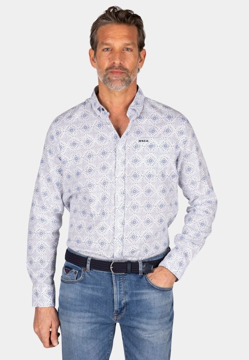 Witte blouse met lange mouwen en een blauw geometrisch patroon, met een knoopkraag en een logo op de borst; gecombineerd met lichtblauwe jeans en een donker gewatteerde riem.