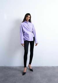 Kvinde iført en oversized lyslilla skjorte med knapper, sorte skinny bukser og sorte spidse hæle, stående op ad en hvid væg.