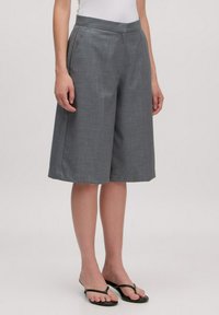 Vrouw in hoge taille grijze wijde knielange shorts, gecombineerd met zwarte slipper sandaaltjes, tegen een effen achtergrond.