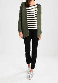Cárdigan de punto verde olivo encima de una camiseta de rayas en blanco y negro de mangas largas, combinado con pantalones negros y zapatos tipo mocasín plateados.