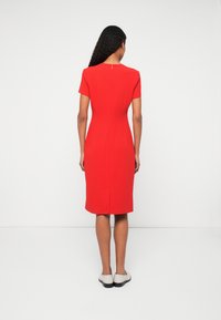 Piros térd hosszúságú ruha, mely testhez álló szabással rendelkezik, rövid ujjakkal és hátsó cipzárral. A szegőnél finom hasítékkal van ellátva a könnyebb mozgás érdekében.