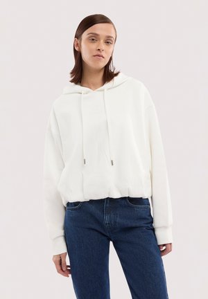 Junge Person trägt einen weißen Hoodie mit Kordeln und dunkelblaue Jeans und steht vor einem schlichten, hellen Hintergrund.