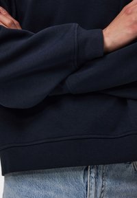 Άτομο που φοράει σκούρο μπλε φούτερ με σταυρωμένα χέρια στο στήθος και ανοιχτό μπλε τζιν τζιν εμφανή κάτω από το φούτερ.