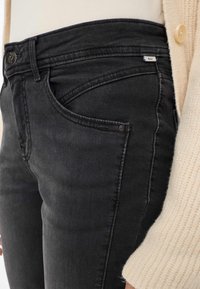 Jeans in denim neri con taglio slim, dotati di tasche e etichetta con marchio. Abbinati a un maglione lavorato a maglia di colore chiaro.