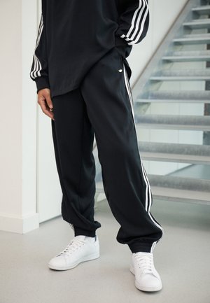 Osoba ubrana w czarne spodnie dresowe Adidas i dopasowaną czarną bluzę z długim rękawem z białymi paskami, stojąca w pomieszczeniu obok metalowych schodów.