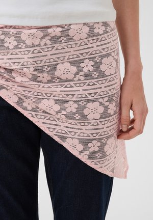 Person trägt dunkle Hosen und ein weißes Oberteil mit einem pinkfarbenen floralen Spitzenasymmetrischen Wickel um die Taille, Hand ruht an der Seite.