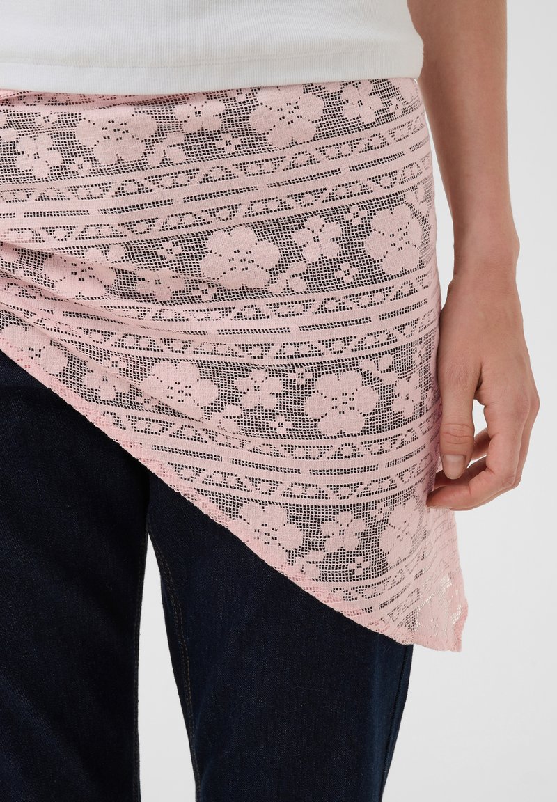 Personne portant un pantalon foncé et un haut blanc avec un drapé asymétrique en dentelle florale rose autour de la taille, main reposant sur le côté.