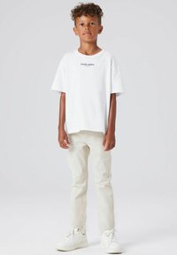 Hvit t-skjorte med korte ermer og teksten "Studio edition", kombinert med lyse beige jeans og hvite joggesko. Enkel design, avslappet passform.