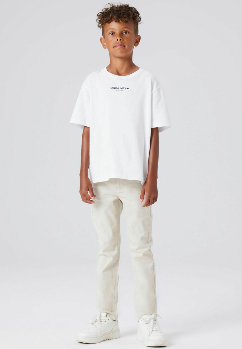 Hvit t-skjorte med korte ermer og teksten "Studio edition", kombinert med lyse beige jeans og hvite joggesko. Enkel design, avslappet passform.