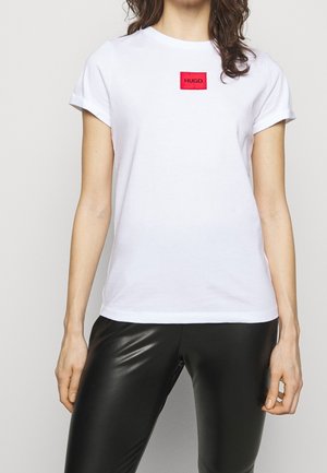Kvinde iført en hvid kortærmet t-shirt med et rødt HUGO-logo-patch og sorte læderbukser, stående mod en ensfarvet baggrund.