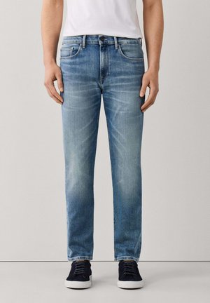 Hombre con vaqueros de pierna recta, descoloridos y de color azul claro, combinados con zapatillas negras y una camisa blanca, de pie frente a un fondo liso.