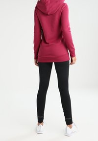 Vínová mikina s kapucí a volnějším střihem, spolu s černými legínami s texturovaným vzorem a reflexními akcenty. Viditelné tenisky.
