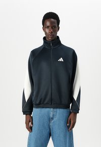 Casaco azul-marinho com fecho éclair e gola alta, acabamentos brancos nas mangas, cintura elástica e o logótipo da Adidas no lado esquerdo do peito. Tecido com uma textura suave.