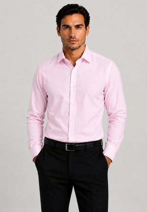 Homme aux cheveux foncés portant une chemise rose clair à manches longues boutonnée, rentrée dans un pantalon noir avec une ceinture noire, mains dans les poches.
